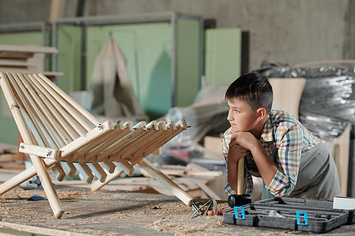 Pensive teenage carpenter leaning on hammer while looking at his finished wooden chair in workshop