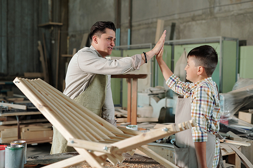 Satisfied male carpenter in apron giving five to teenage son when they have finished wooden chair in workshop