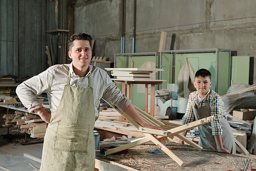 Portrait of satisfied furniture makers in aprons standing at desk with wooden chair and shavings in workshop, family business