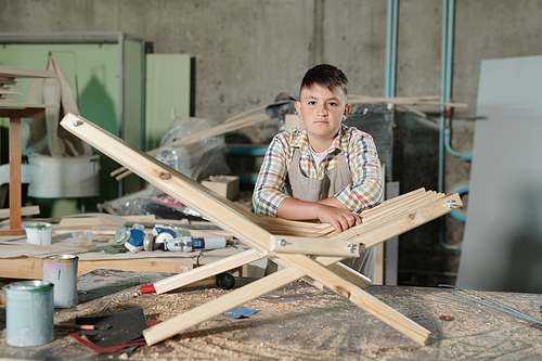 Portrait of serious teenage boy standing at table with his first wooden chair in carpentry shop