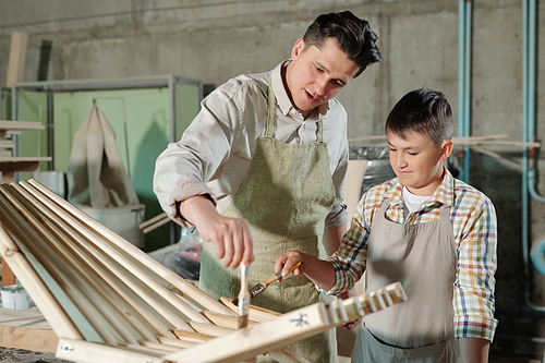 Busy middle-aged father in apron and his teenage son applying varnish to wood while making wooden chair in carpentry workshop