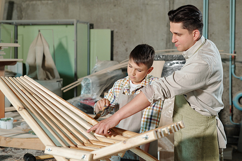 Father pointing son at unvarnished places of wooden chair to son while they making furniture in workshop