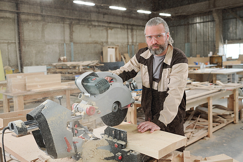 Confident aged professional in eyeglasses and workwear standing by workplace while cutting thick wooden board with electric saw