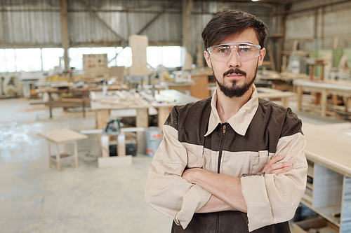 Young serious worker or engineer of furniture producing factory in uniform and eyeglasses standing in front of camera in large workshop