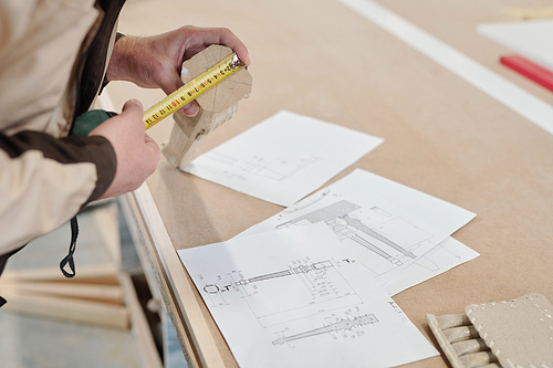 Modern factory worker measuring wooden part of furniture while holding it over sketches on table and comparing length indication