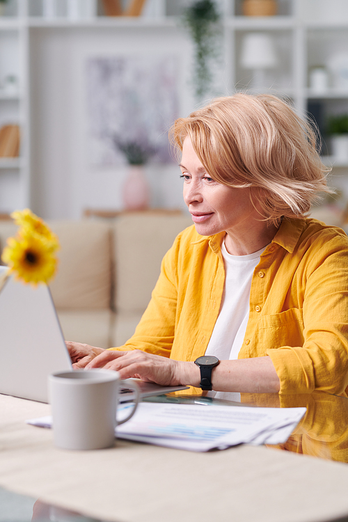 Pretty blond mature female in yellow jacket sitting by desk in front of laptop while networking in home environment