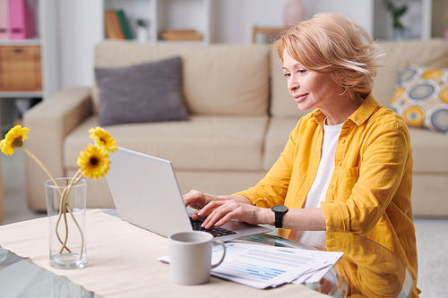 Happy blond mature female in casualwear sitting by desk in front of laptop and working remotely in home environment during quarantine