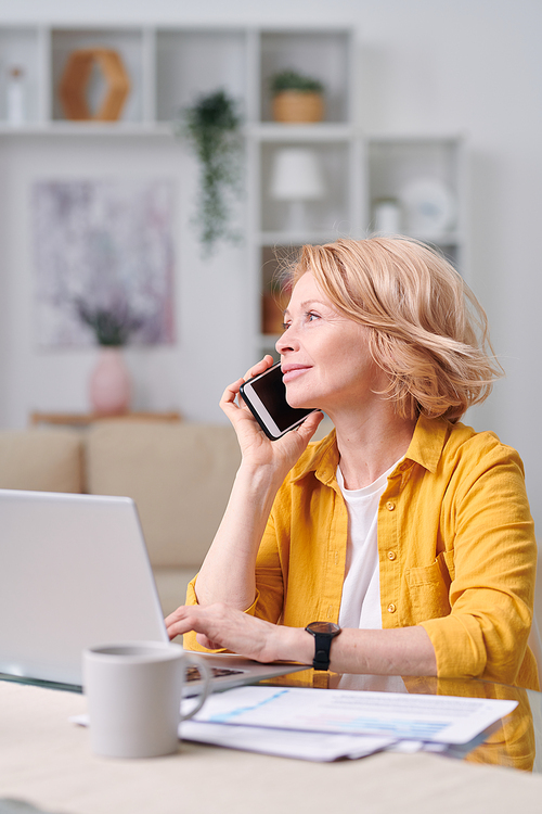 Successful mature female agent with smartphone calling one of clients while sitting by workplace in home environment