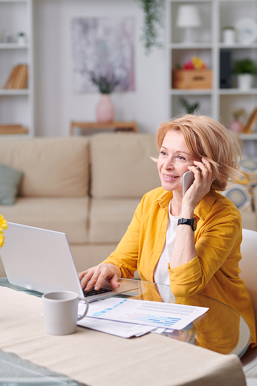 Mature cheerful businesswoman with blond hair talking to someone on smartphone while working remotely at home during quarantine