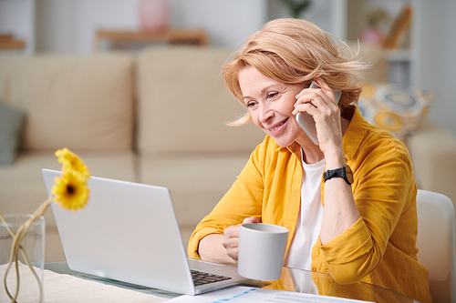 Happy mature businesswoman with mug and mobile phone consulting one of clients while working remotely during home isolation