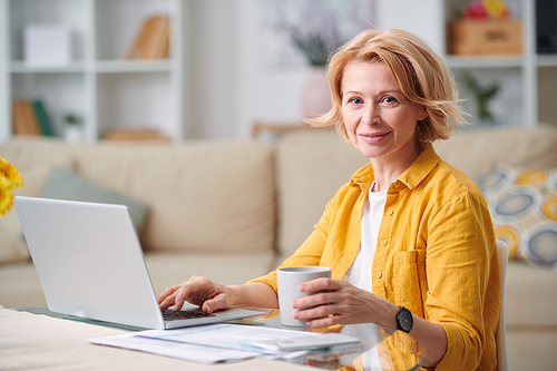 Successful mature businesswoman with mug of tea looking at you while sitting by desk in front of laptop at home during quarantine