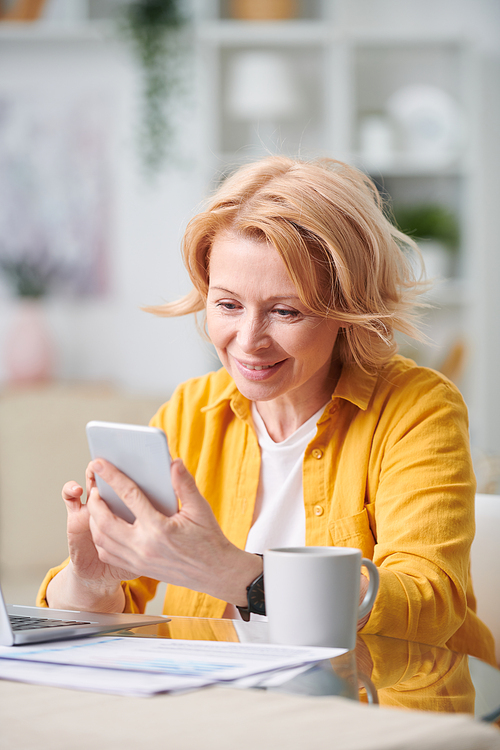 Happy blond woman with smartphone looking through working plan while sitting by desk in home environment during quarantine