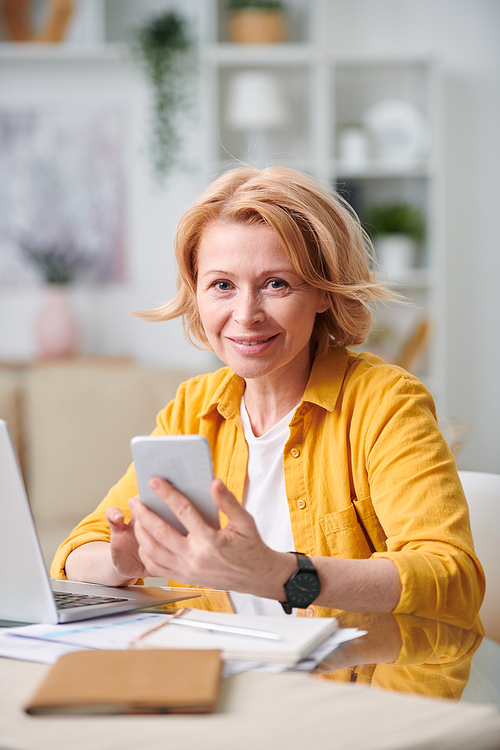 Busy mature female with blond hair looking at you while scrolling in smartphone in front of laptop in home environment
