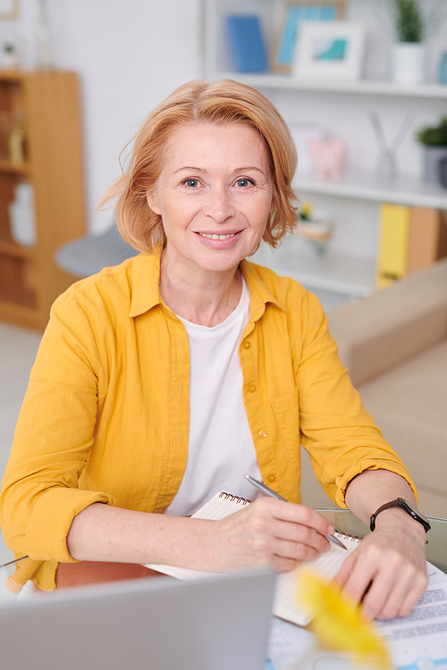Blond mature female in white t-shirt and yellow shirt making notes in notepad while working at home during quarantine