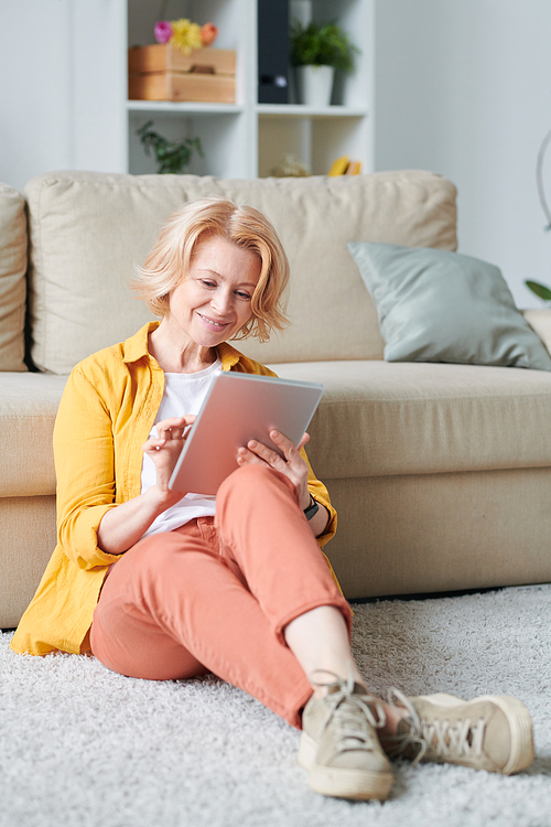 Mature blond relaxed businesswoman with mobile gadget sitting on the floor by couch in living-room while planning work during quarantine