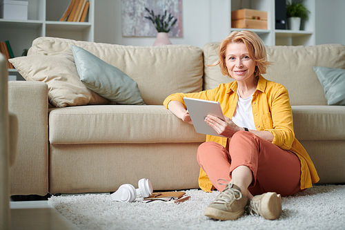 Cheerful mature businesswoman in casualwear sitting by couch in living-room and browsing in the net while working remotely