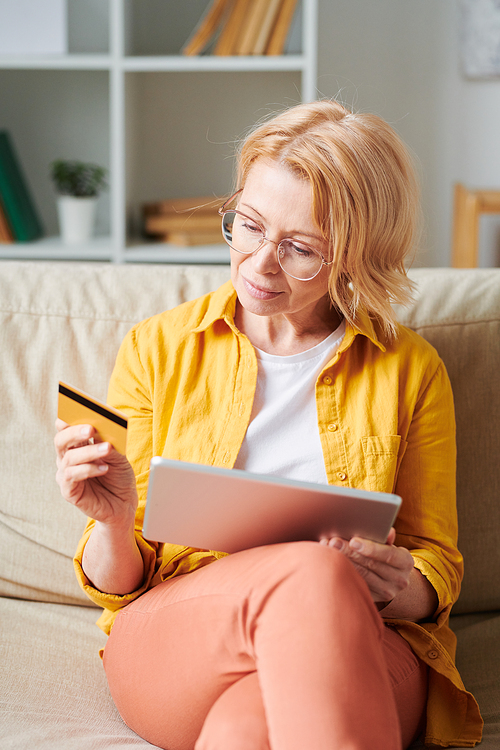 Blond mature female with touchpad and credit card sitting on couch after working day and shopping through internet in living-room