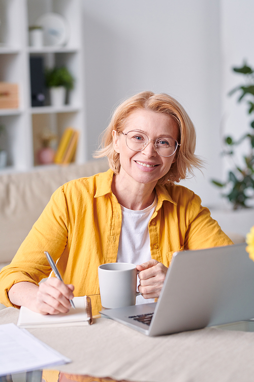 Pretty smiling blond businesswoman in casualwear and eyeglasses looking at you while working at home during self isolation
