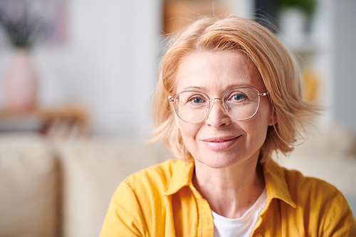 Pretty smiling mature female in eyeglasses and casualwear standing in front of camera and looking at you in home environment