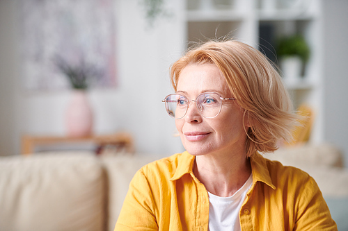 Contemporary blond mature female in yellow casualwear and eyeglasses expressing serenity while having rest in home environment