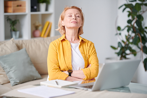 Serene mature blond woman in casualwear relaxing on couch in front of laptop with her eyes closed during remote work at home