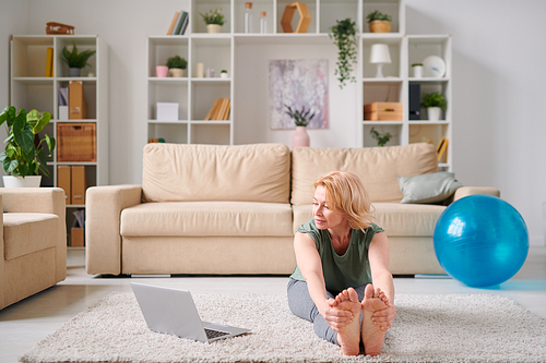 Mature active woman in sportswear sitting on the floor of living-room in front of laptop and doing fitness exercises in the morning