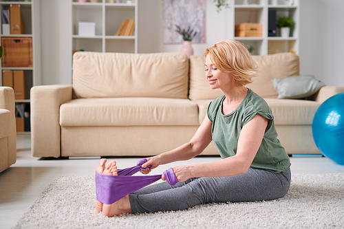 Happy mature woman in grey leggins and t-shirt sitting on the floor of living-room and doing exercise for legs during home quarantine