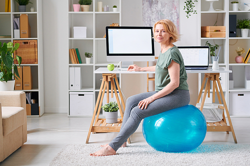 Blond mature female in activewear sitting on fitball by desk in front of computer monitor while going to exercise at home