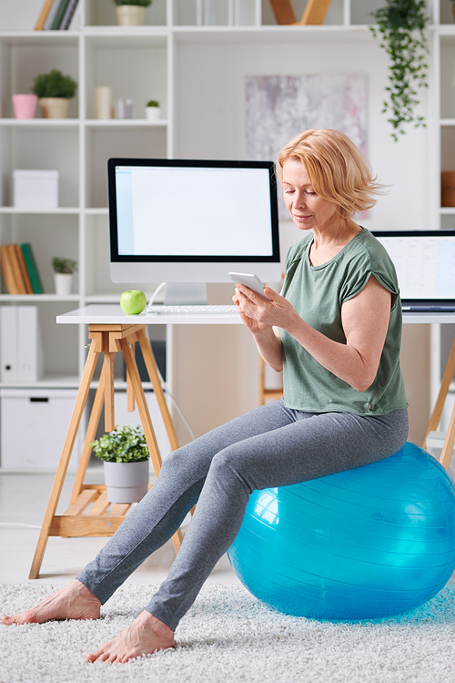 Pretty mature woman in activewear sitting on fitness ball and scrolling through online training course in smartphone at home
