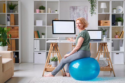 Mature sportswoman in activewear looking at you while sitting on fitball in front of computer screen and surfing for online workout