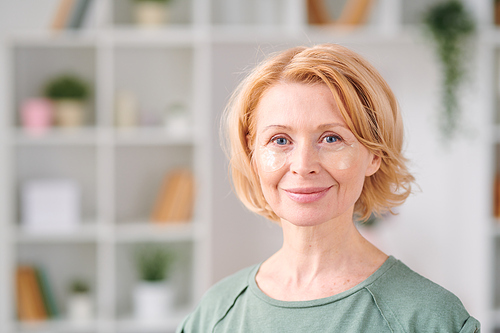 Smiling mature woman with under eye patches looking at you while standing in front of camera on background of shelves with flowerpots and books