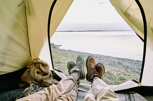 Legs of young amorous couple in casualwear lying inside open tent in front of seaside and relaxing while hiking on summer weekend