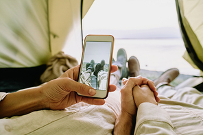 Close-up of unrecognizable couple of hikers relaxing in tent and taking photo of crossed legs against lake