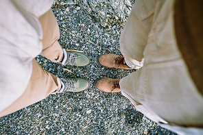 Overview of legs in pants and feet in boots of young traveling couple standing on riverbank on summer day during their journey