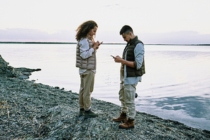 Concentrated young couple standing on pebble beach and using gadgets while checking connection
