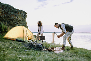 Young male traveler with ax chopping wood on riverbank covered by green grass on background of his wife and their tent