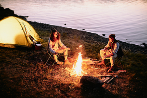 Young romantic couple in casualwear sitting by campfire on riverbank, talking and frying sausages or marshmallows by their tent