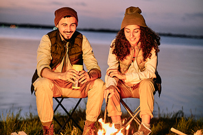 Cheerful young couple in hats sitting on folding chairs near bonfire and chatting together at night