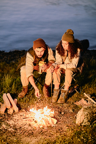 Young hikers sitting by campfire in the evening on background of river or lake, relaxing and having tea or coffee with fried marshmallows