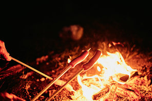 Close-up of unrecognizable couple frying sausages on sticks above camping fire at night
