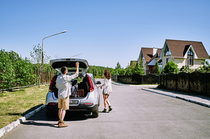 Young man closing car trunk with backpacks and bags while his wife opening door of their vehicle standing on road in countryside