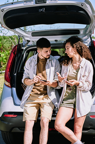 Young restful couple in casualwear sitting on car trunk and discussing route of their journey while going for hike on summer weekend