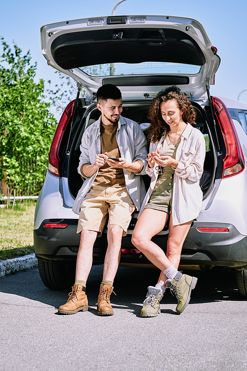 Positive active young hikers in boots leaning on car trunk and discussing hiking route using phone apps