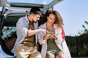 Young couple leaning on car trunk and using smartphone while checking hiking list before trip