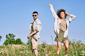 Joyful young woman dancing in green grass against blue sky and expressing gladness while her husband standing near by