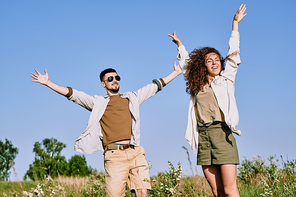 Cheerful excited young couple in comfortable wear standing with raised arms while enjoying travel in country