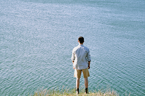 Rear view of young male traveler in shirt and shorts standing on riverbank close to water and enjoying nature and solitude on summer day
