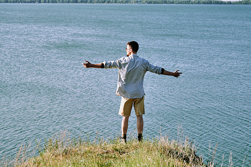 Rear view of young man in shirt standing with outstretched arms on edge of cliff and contemplating beautiful waterscape