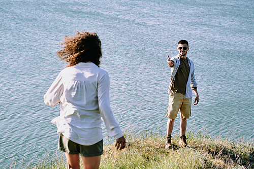 Smiling young male hiker stretching arm to his wife and inviting her to come closer and enjoy wonderful view of lake on sunny day