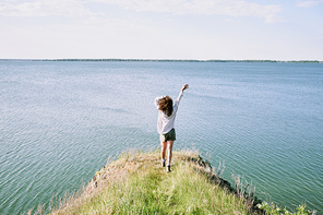 Back view of young ecstatic woman with dark curly hair enjoying freedom and sunny day while moving towards water with her arms raised